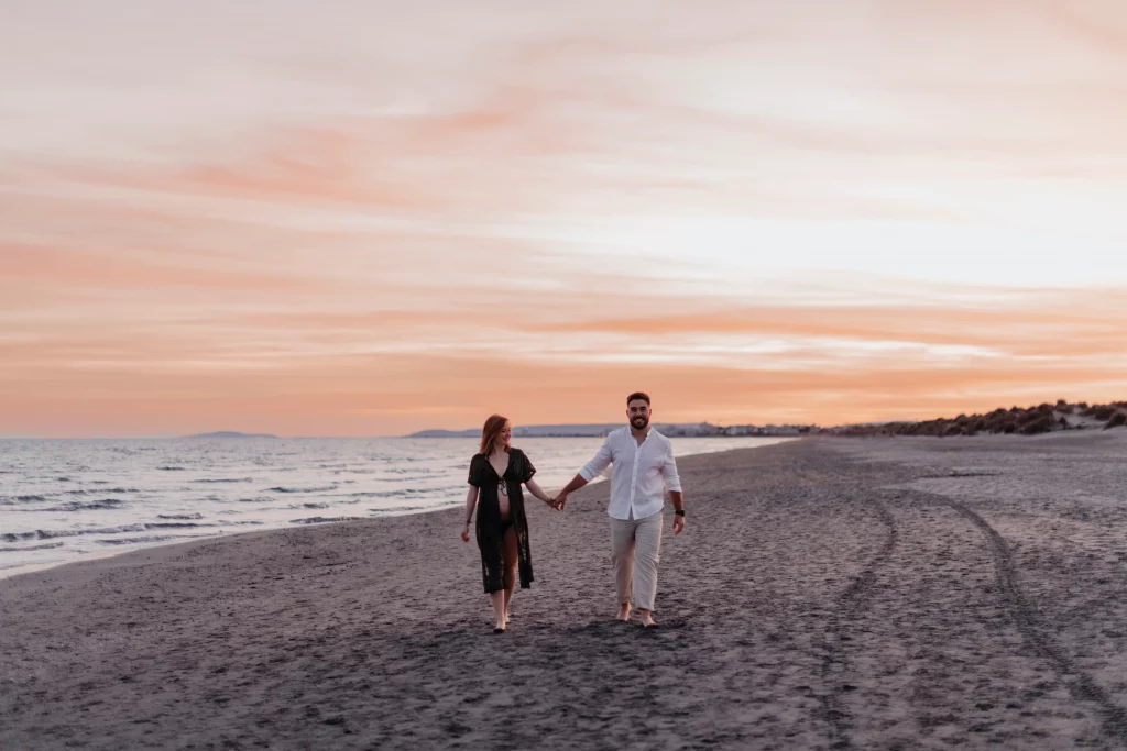 Couple de futurs parents se baladant main dans la main sur la plage.